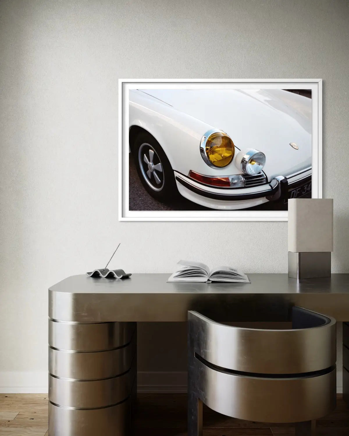 A framed art print of a white classic Porsche car, shown hanging on a light gray wall above a modern metallic desk and chair.
